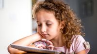 A young girl with curly, light brown, golden hair and a pink shirt is sitting, resting her arm on a yellow couch, reading on a tablet in her house.
