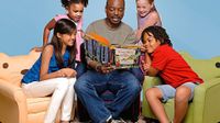 LeVar Burton, associated with Reading Rainbow, is sitting on a blue couch chair reading a book to four kids. A girl is sitting on a cat chair to his left, a boy is sitting on a frog chair to his right, and two girls are standing behind them.