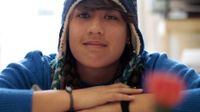 A closeup of a teenage student in a blue sweater and beanie sitting, arms folded over a book.