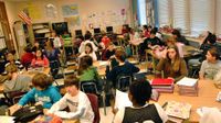 Students are sitting in a classroom at rectangular and circular tables stacked with piles of notebooks and textbooks.