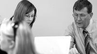 A male teacher is sitting at a desk, looking at a laptop. A female teacher is standing beside him, looking at the laptop, talking.