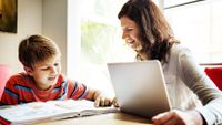 A female adult with a laptop in front of her is smiling, sitting at a table next to a young boy who is looking at an opened book.