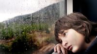 Boy leaning his head against a window sill looking out at the rain