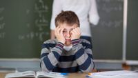 A middle school male student sits at his desk, frowning and holding his head in his hands.