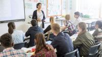 A high school teacher stands before her students and speaks while they listen.