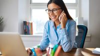A young female teacher wearing glasses is at her desk smiling and on the phone.