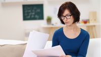 A female teacher in a blue shirt and glasses is sitting in a classroom alone, looking at papers.