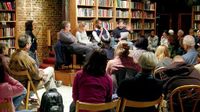 Room of seated adults surrounded by book shelves with four people front and center speaking