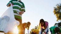 A group of young teens are outside in the sun picking up garbage from the grass and putting it into white trash bags.