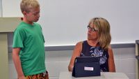 A female teacher in glasses is sitting at a desk with a tablet in front of her looking up towards a young boy. The young boy is wearing a green shirt and is standing to her left.