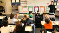 Teacher in front of class pointing to students sitting at desks with raised hands