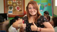 Girl in a classroom standing in front of students working giving two thumbs up