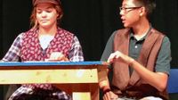 Girl wearing a cap and a boy sitting at a table on stage