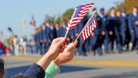 Picture of two people waving small U.S. flags with a memorial day parade in the background