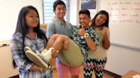 Three teenage students in a classroom, standing side by side, are holding another student and giving a thumbs up.