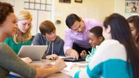 A male teacher assists several students who are smiling and working on their laptops.