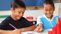 Two elementary school boys sit together, writing and laughing.