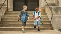 Two young boys wearing backpacks rushing down the front steps of school
