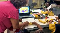 Woman doing a project with young kids using bananas and Play-Doh with a laptop in the center of the table open to an app