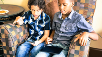 A photo of 2 young boys sitting in a big easy chair, playing games on their tablets.