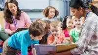 A teacher shares a book with students as another teacher looks on.