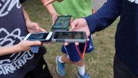Three students are standing on the grass, a close up on their arms extended, smartphones in their hands, and Pokemon Go on their screens.