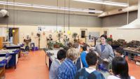 A male teacher wearing goggles and a long jacket in a large science laboratory is surrounded by a group of eight students, all looking up at him, also wearing protective goggles.