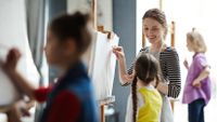 An adult guides several young children as they stand at easels.