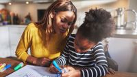 Seated at a kitchen table, a female adult assists a young female child with a writing assignment.