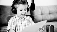 A black and white image of a young boy wearing headphones, reading from papers, speaking into a hanging microphone