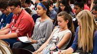 A large group of students are sitting in chairs, close together, with pens and paper out, writing.