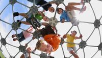 Eight young students are outside on top of a metal, geodesic dome at a playground, looking down.