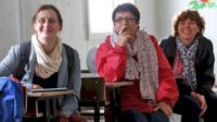 Three women are sitting at desks, looking at the front of the classroom, smiling.