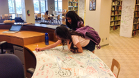 A photo of 2 female students writing a whiteboard table with colored markers.