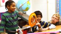 Boy showing his project to classmates -- a silver tube over his arm with a globe tied to the top and a fan at the end of the tube