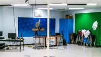 End of a room set up with computers, green screen, and a broadcast area with school logo as a backdrop
