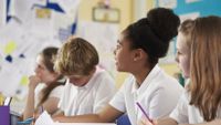 Four middle-school aged students sit in a row, writing and facing the board.