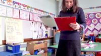 Teacher standing alone in classroom reading off a clipboard