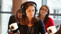 Woman sitting on a panel in front of a microphone wearing headphones with young people sitting behind her