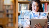 A female student looks at a tablet computer with holographic images of data charts floating around it.