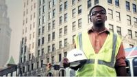 Boy wearing a yellow safety vest holding his hard hat, standing on a busy street in front of a very large building