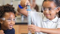 Two young students wearing safety goggles; one pouring liquid into a beaker