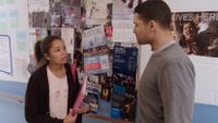 A female teen is standing in a school hallway talking to a male teacher; there are dance fliers on a bulletin board on the wall beside them.