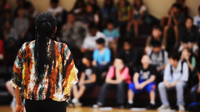 A photo of a woman talking to a large group of students in a school auditorium.