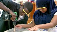 Wearing safety goggles, a boy working a drill while a girl steadies the project using two hands