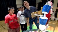 Two boys and a mentor student watching water pour into brightly painted plastic cups causing the wheel to turn