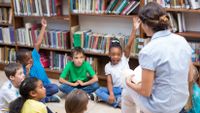 A teacher sits on a chair and reads to students sitting on the floor.