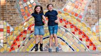 Two girls jumping up in front of a large tiled wall of a sun