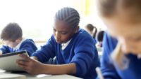 Students in uniform sweaters sit in a classroom and write by hand.