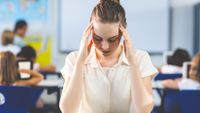 A teacher in class rubs her temples and looks down at her desk.